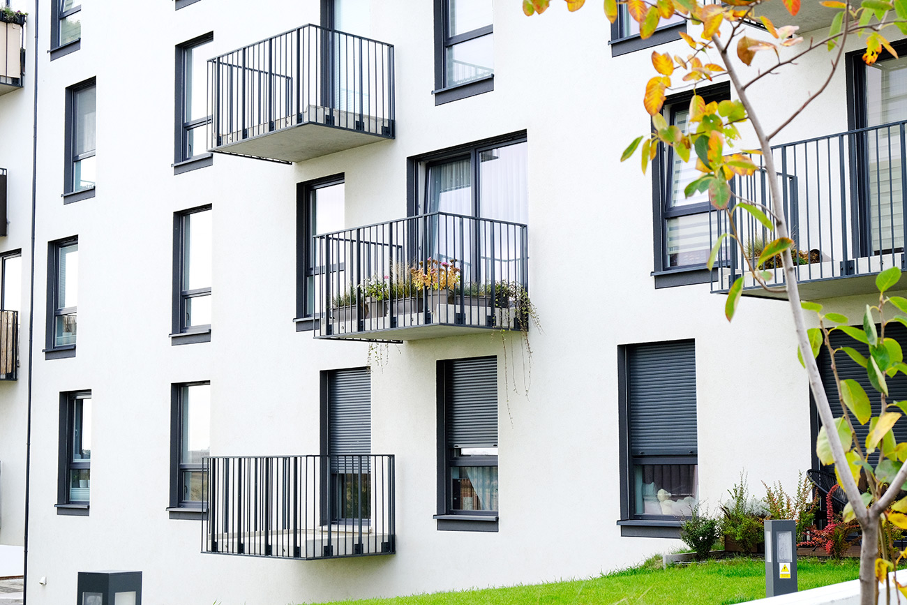 Mehrstöckiges Wohngebäude mit weißen Fassaden und Balkonen mit schwarzen Geländern, im Vordergrund grüner Rasen und ein Baum mit gelben Blättern