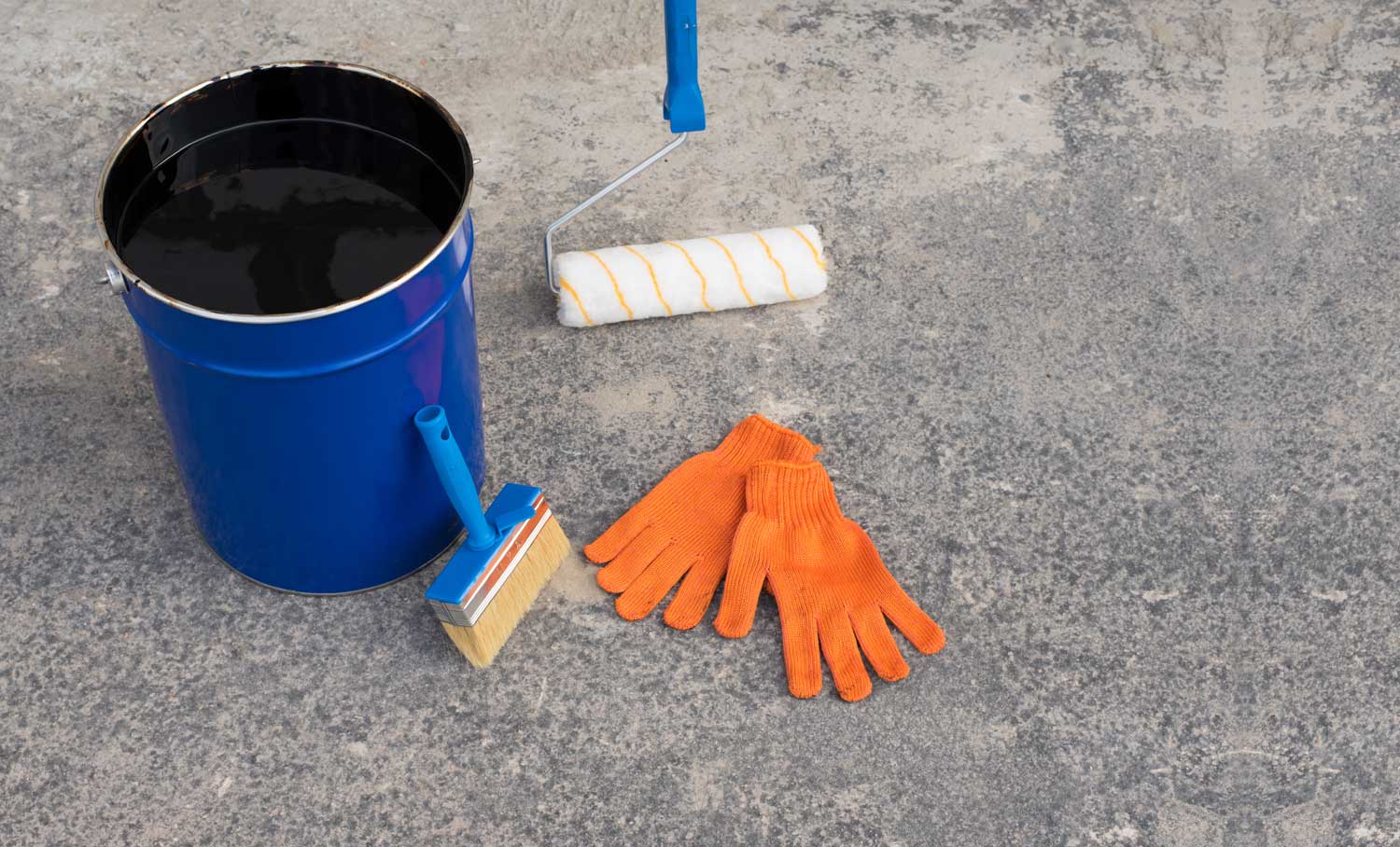 Blauer Farbeimer mit schwarzer Flüssigkeit, Farbroller mit weißem Bezug und orangene Arbeitshandschuhe auf grauem Boden