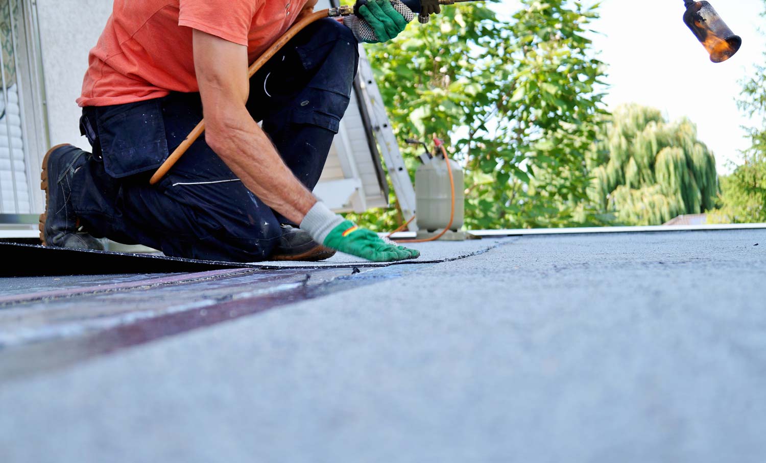 Handwerker kniet auf Dach und befestigt Dachpappe mit Hammer und Handschuhen