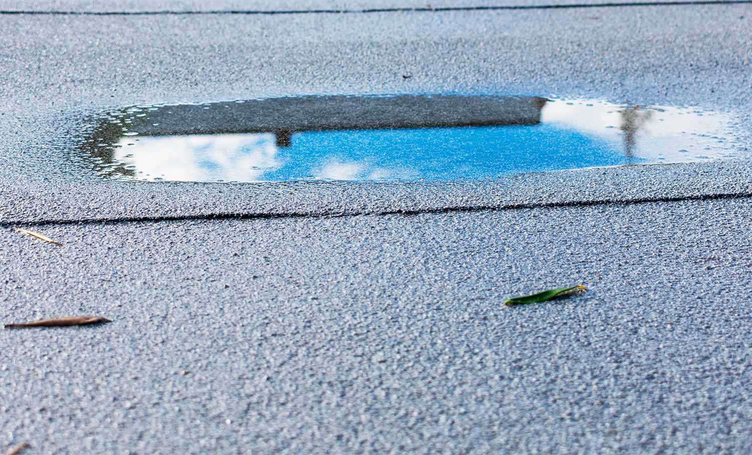 Wasserpfütze auf Bittumenmatte auf einem Flachdach mit Spiegelung des blauen Himmels und Wolken